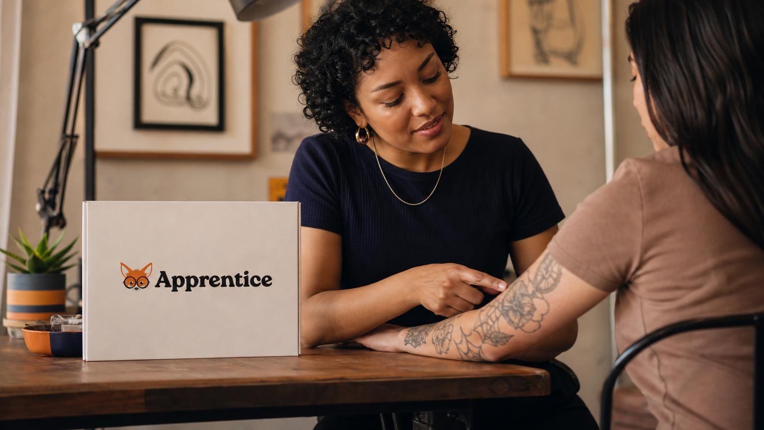 A woman examines a floral tattoo on another person's arm while sitting at a wooden table next to an "Apprentice" branded box.